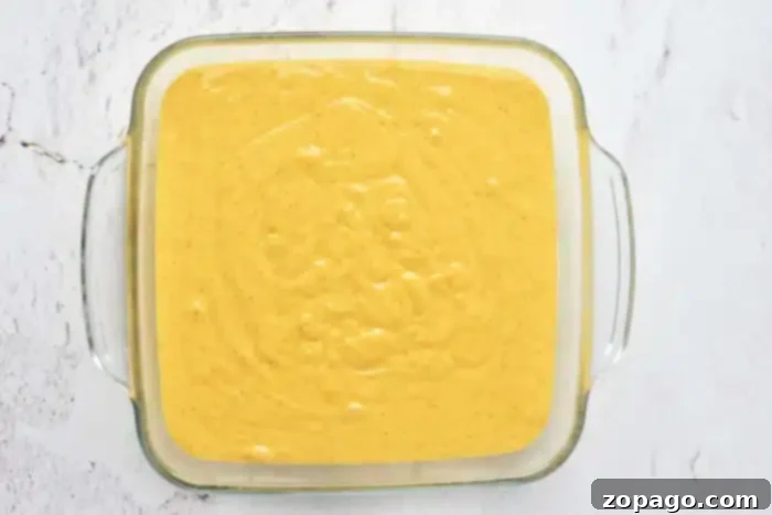 A square glass baking dish holds a smooth, golden-yellow batter, ready for baking into a pumpkin cake with yellow cake mix. A few small air bubbles dot the surface, indicating its freshly mixed state, all set on a light-colored marbled countertop.