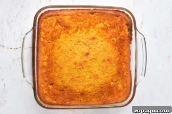 A square glass baking dish presents a freshly baked, golden-brown pumpkin cake with yellow cake mix, cooling on a light-colored textured surface. The cake's edges are perfectly crisp and slightly darker, with a smooth, even top, indicating ideal doneness.