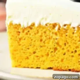 A close-up of a slice of pumpkin cake with yellow cake mix, featuring a thick layer of white frosting. The cake has a yellow, airy interior with visible bubbles and is placed on a beige plate.