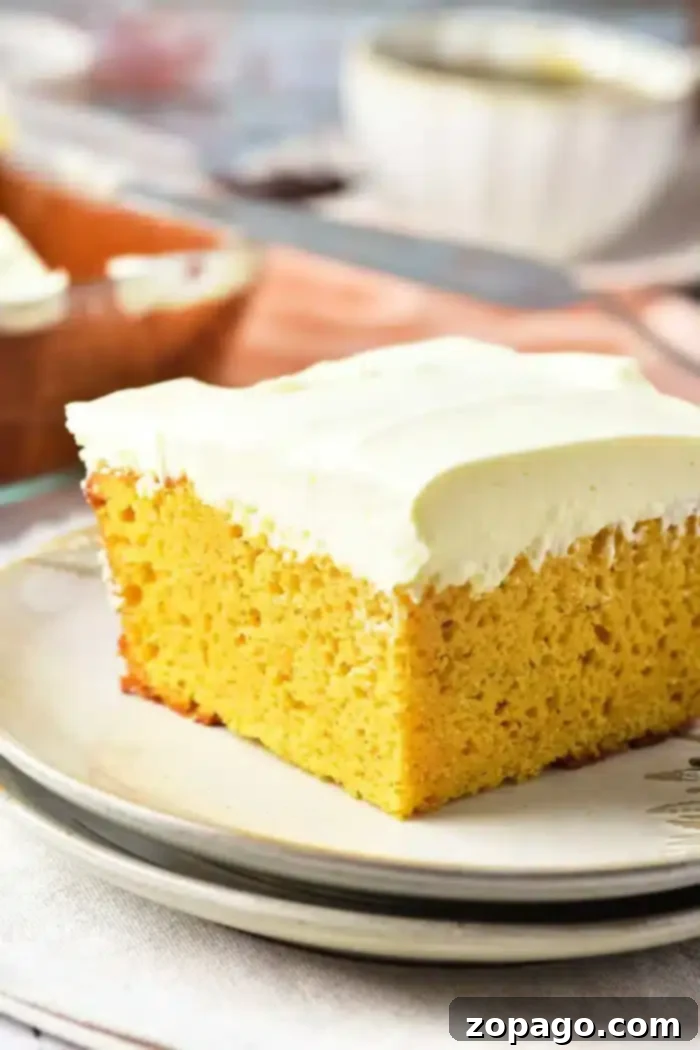 A beautiful close-up of a slice of yellow pumpkin cake, adorned with a thick layer of smooth white cream cheese frosting, presented on a plate. In the blurred background, more frosted cake, a knife, and a bowl suggest a freshly prepared dessert table, set on a light-colored tablecloth.