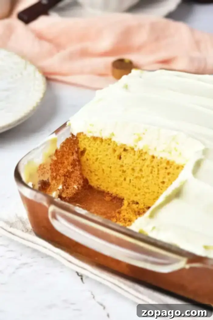 A rectangular glass baking dish holds a generously frosted pumpkin cake made with yellow cake mix. A square piece has been neatly removed, showcasing the cake's moist interior and the creamy white frosting. The dish rests on a light-colored kitchen cloth, with a serving plate and a peach-colored fabric artfully arranged in the background.