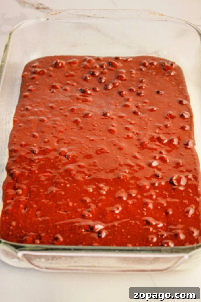 Brownie Cookie Dough Bars 3 A clear glass baking dish filled with thick, dark chocolate brownie batter, resembling the foundational layer for crafting brookies. The smooth batter, with small, uneven bubbles, is spread evenly and looks ready for the next layer on a light-colored countertop.