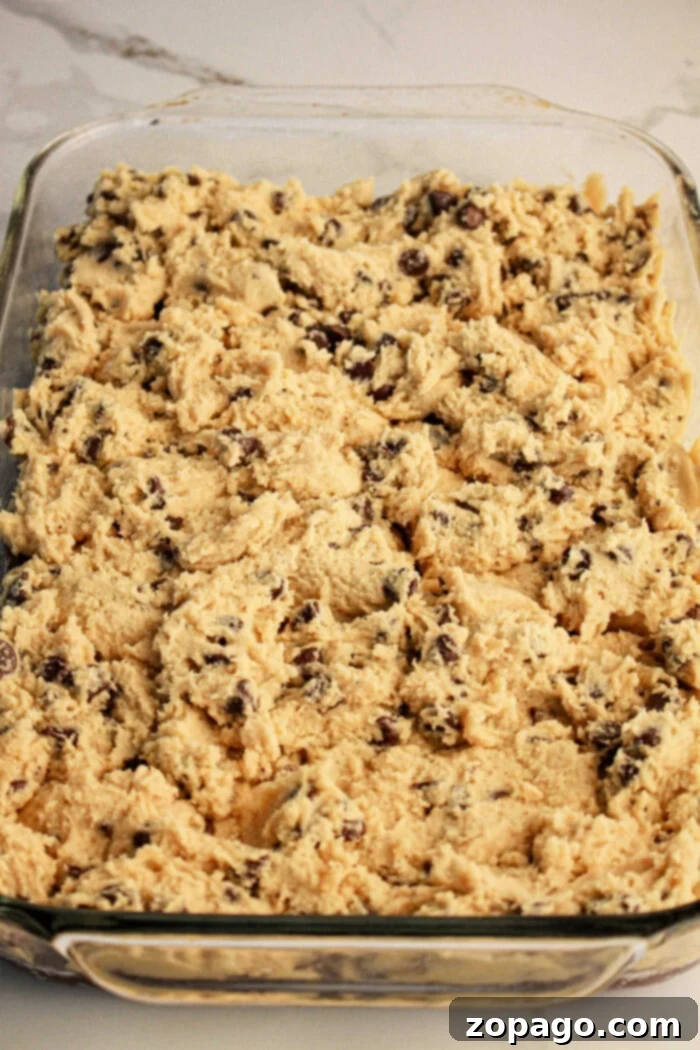 Fudgy Cookie Dough Brookies 4 Close-up of a glass baking dish filled with unbaked chocolate chip cookie dough, spread evenly like an easy step in learning how to make brookies with brownie mix and cookie dough. The dough is light brown with visible chocolate chips, ready for baking.