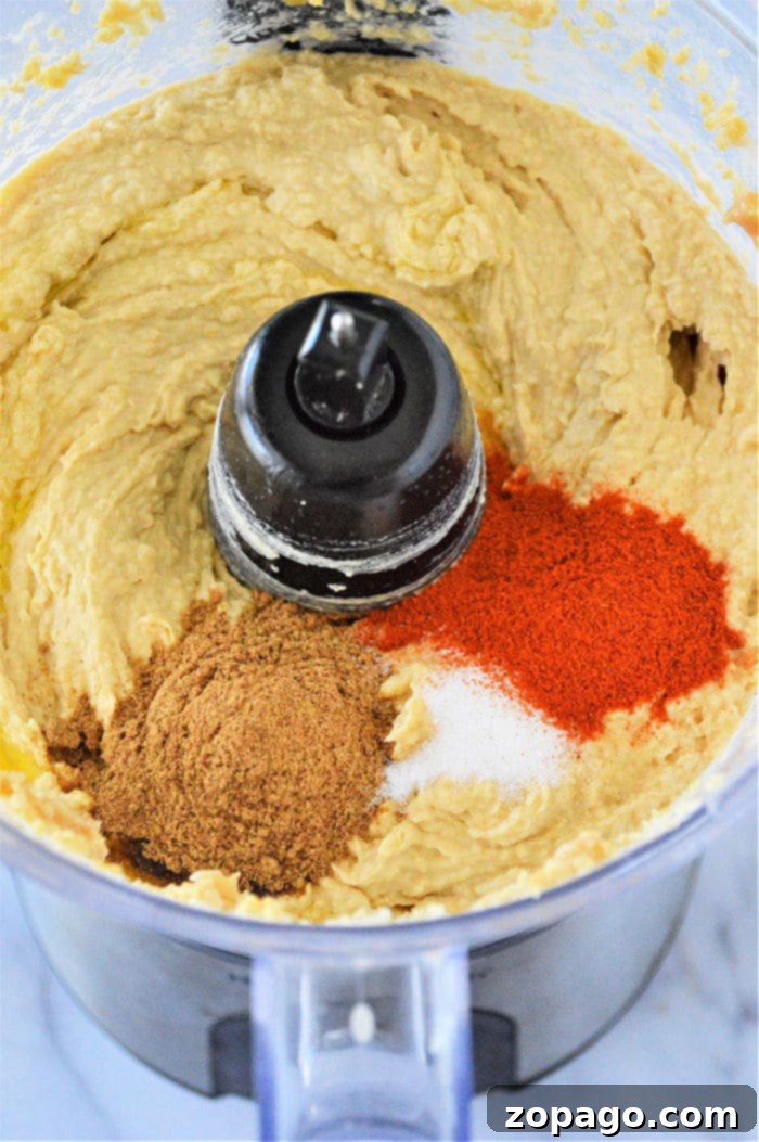 Make creamy, easy hummus at home with canned chickpeas. Add garlic or other flavors for a delicious, healthy, gluten-free snack. A close-up shot of creamy garlic hummus garnished with paprika and fresh herbs.