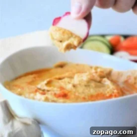 Effortless Hummus: Canned Chickpea Edition 7 Hand dipping sliced radish into a creamy, easy hummus made with canned chickpeas, with garlic and fresh vegetables in the background.