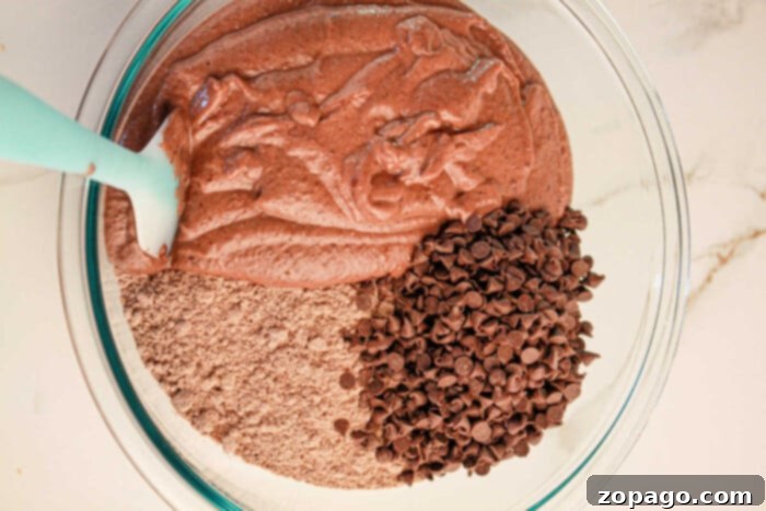 Decadent Black Bean Brownies 4 A bowl of black bean brownies, featuring batter and dry mix studded with chocolate chips, is being expertly combined with a turquoise spatula.