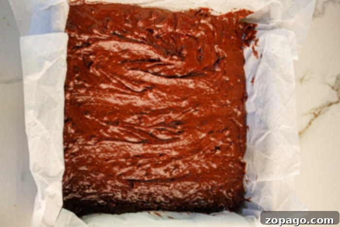 Decadent Black Bean Brownies 5 A parchment-lined square baking tin rests on a marble surface, brimming with rich chocolate black bean brownie batter, ready to mix magic in the oven.