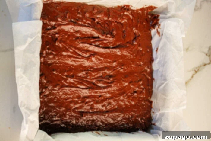 Secretly Fudgy Black Bean Brownies 5 A parchment-lined square baking tin filled with rich, smooth chocolate black bean brownie batter, perfectly prepared and ready to be baked in the oven, resting on a clean marble surface.