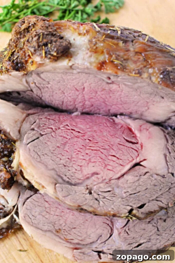 A close-up of a sliced, medium-rare Boneless Rib Roast. The beef is browned on the outside and pink with a slightly red center on the inside. It is garnished with sprigs of fresh herbs in the background. The meat is placed on a wooden cutting board, ready to be served.