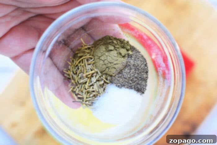 A hand holding a clear glass bowl containing a mix of dried rosemary, ground black pepper, salt, and another greenish-brown spice. Perfect for seasoning your Boneless Rib Roast Recipe, demonstrating the simple yet effective dry rub ingredients. A wooden cutting board is visible in the background.