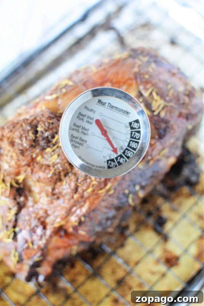 Tender Boneless Rib Perfection 5 A meat thermometer is inserted into a cooked boneless rib roast, showing a temperature reading. The roast, seasoned with herbs, rests on a wire rack set on a baking sheet. The thermometer's dial displays various meat types and corresponding temperature ranges.
