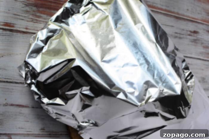 Tender Boneless Rib Perfection 7 A piece of food, possibly involving a boneless rib roast recipe, is wrapped in crinkled aluminum foil on a wooden surface. The aluminum foil reflects light and covers the object completely. The wood surface has a whitewashed finish with visible grain patterns.