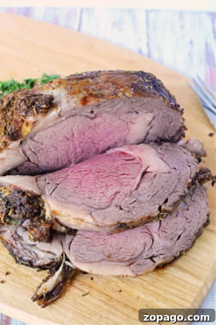 Tender Boneless Rib Perfection 8 A beautifully sliced boneless rib roast on a wooden cutting board showcases medium-rare perfection in the center with a darker, well-done outer layer. Fresh herbs are sprinkled on top, accompanied by green garnish in the background. A fork is partially visible on the right side, completing this exquisite Boneless Rib Roast Recipe.