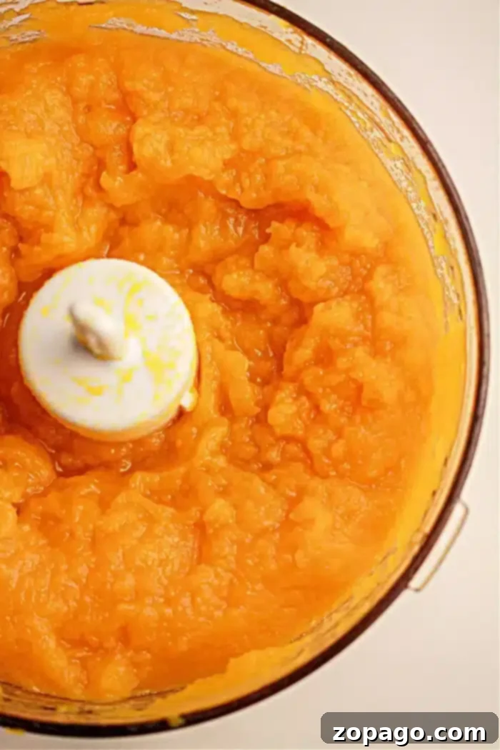 Crockpot Pumpkin Bliss 8 A food processor bowl filled with smooth, bright orange pumpkin puree, perfectly blended and ready for use in various autumn recipes. The white blade of the processor is clearly visible in the center.