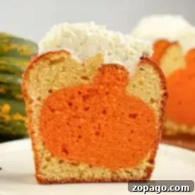 A single piece of cake on a white plate reveals an orange pumpkin shape inside the cake. Topped with a layer of white frosting, the cross-section showcases the charming design. In the blurred background, another piece of cake awaits its turn.