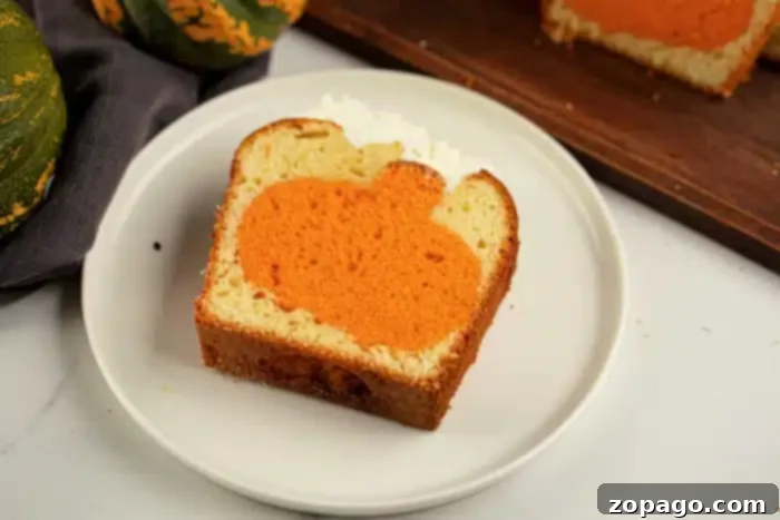 Pumpkin Surprise Loaf Cake 9 A charming slice of baked loaf cake on a white plate features a distinct orange pumpkin shape hidden within. In the background, a wooden cutting board holds another slice of the loaf, accompanied by a festive cloth and decorative green pumpkins, signaling this delightful cake is ready for a fall celebration.