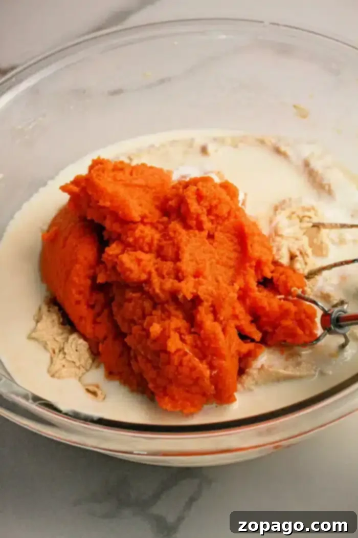 Spiced Pumpkin Poke Cake 3 A glass mixing bowl holds vibrant orange pumpkin puree, alongside pale beige dry ingredients, and a liquid component. An electric mixer's beater is partially submerged, poised to blend these elements into a delightful Pumpkin Poke Cake batter.