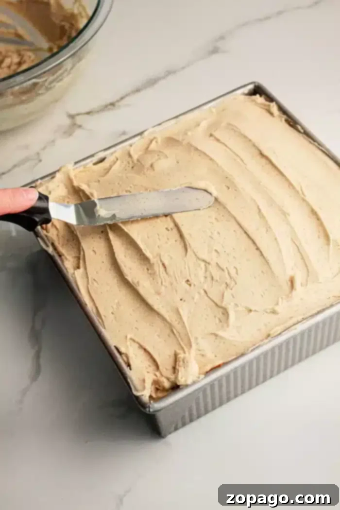 Spiced Pumpkin Poke Cake 5 A hand expertly spreads a smooth, beige-colored frosting across the top of a square Pumpkin Poke Cake, still nestled within its metal baking pan. An icing spatula is used to achieve an even layer, with a glass bowl containing additional frosting visible in the background on a marble countertop, ready for more delectable finishing touches.