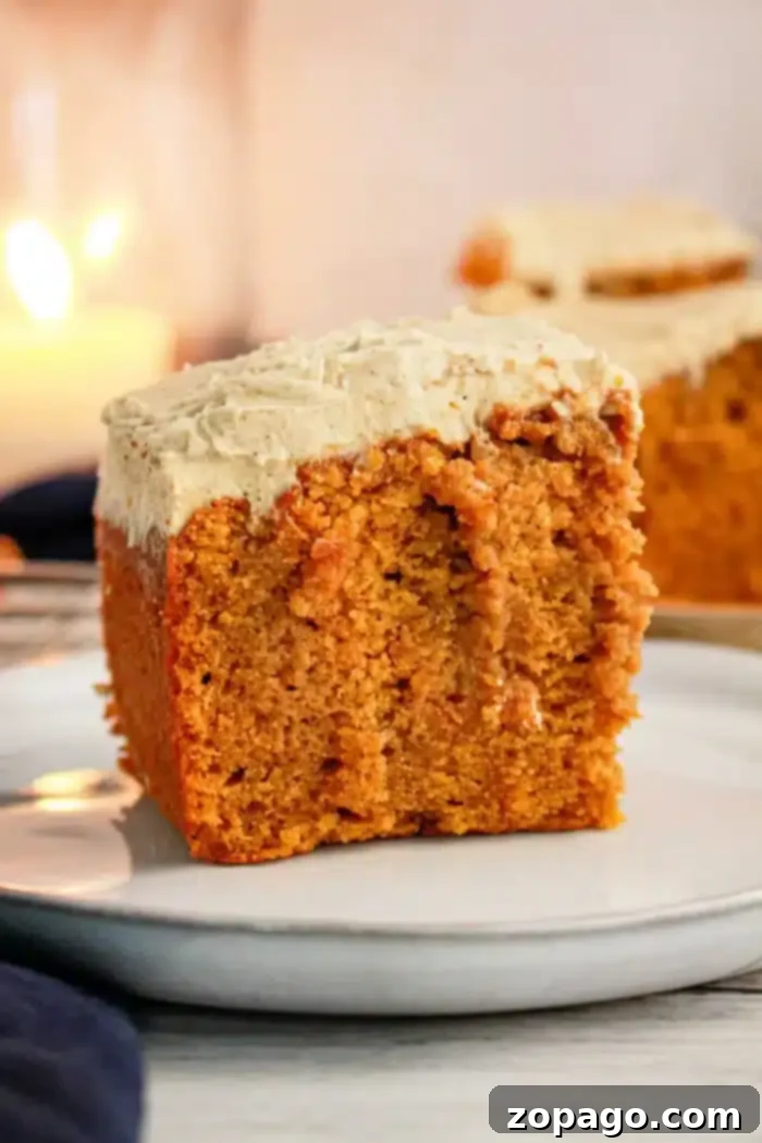 Spiced Pumpkin Poke Cake 6 A close-up of a slice of Pumpkin Poke Cake with cream cheese frosting on a white plate. The cake has a golden-brown color and a moist texture, while the thick frosting covers the top of the slice. A blurred background shows another slice and a lit candle.