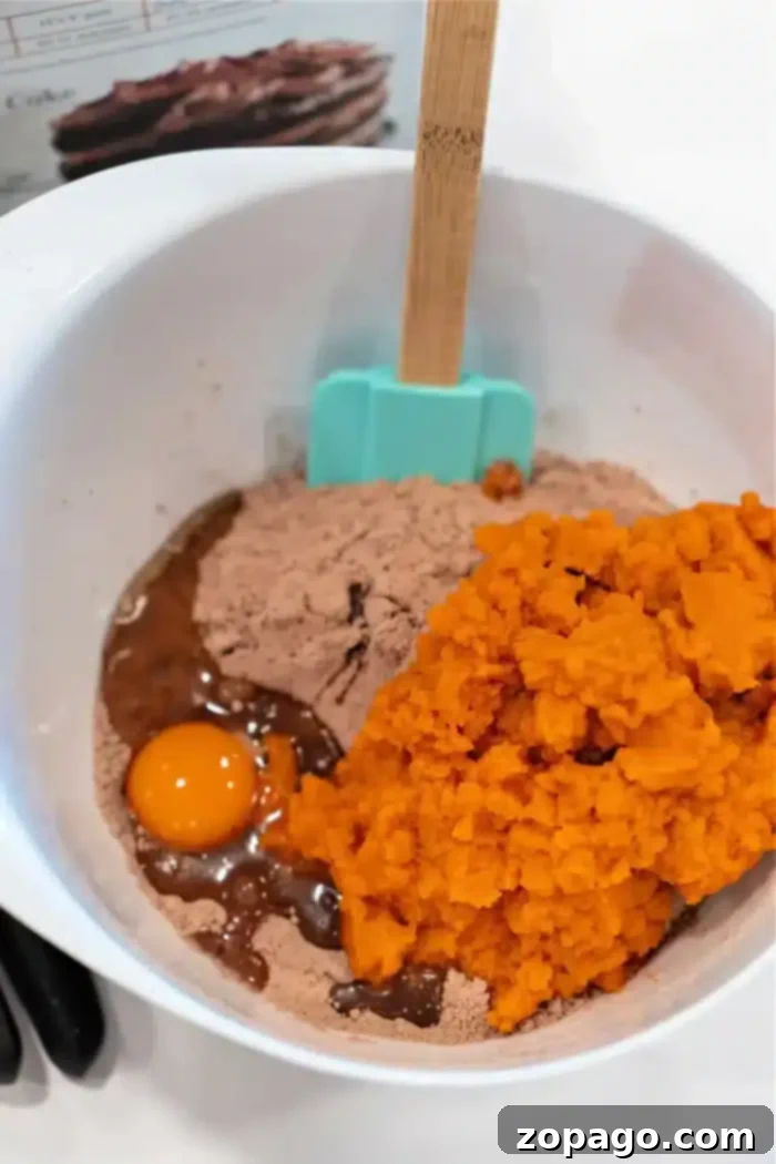 Pumpkin Swirl Boxed Brownies 4 A white mixing bowl contains ingredients for pumpkin brownies. Visible items include a raw egg, vibrant pumpkin puree, and rich brown cake mix. A blue spatula and a wooden one are also in the bowl, with a recipe card slightly visible in the background.