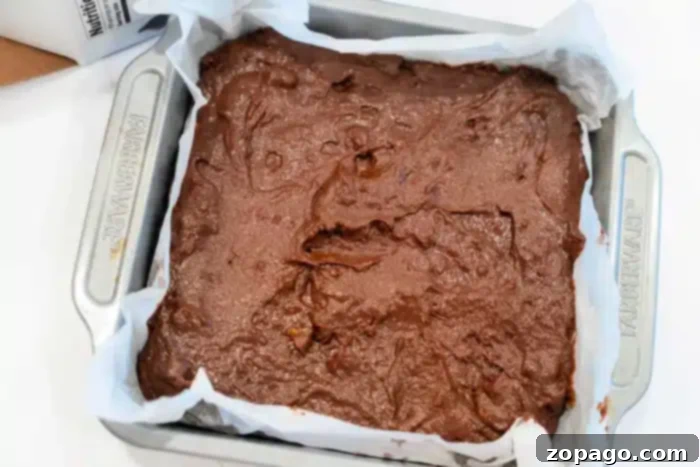 Pumpkin Swirl Boxed Brownies 5 A square baking dish with parchment paper is filled with unbaked pumpkin brownie batter. The batter is spread unevenly across the surface, ready for baking. The pan is labeled "Farberware.