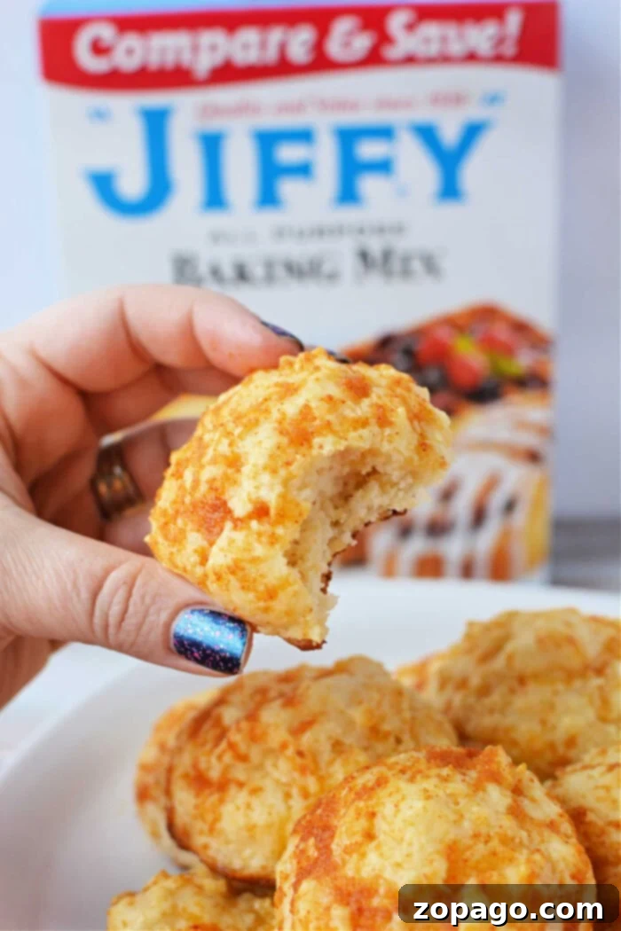 Easy Cheesy Jiffy Biscuits 2 A hand holds a bitten cheddar biscuit, with a Jiffy Mix box adding a tantalizing promise of more flaky treats in the background.