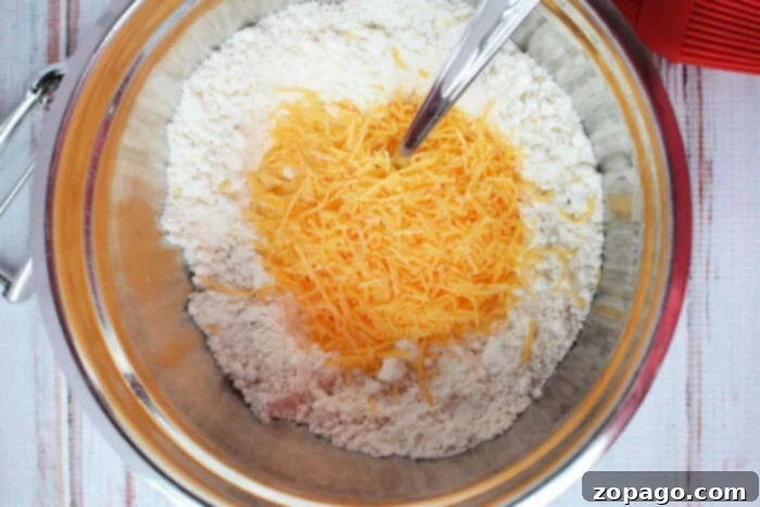 Easy Cheesy Jiffy Biscuits 4 A mixing bowl filled with flour, shredded cheddar cheese, and a spoon rests on a wooden surface, resembling the perfect start for savory biscuits using Jiffy Mix.