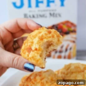 A hand holds a bitten cheddar biscuit, with a Jiffy Mix box adding a tantalizing promise of more flaky treats in the background.