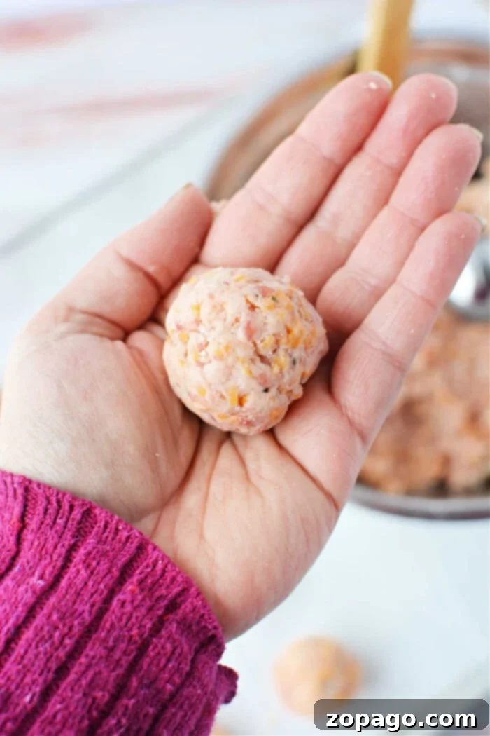 Red Lobster Biscuit Sausage Poppers 5 A hand wearing a pink sweater holds a perfectly formed, raw sausage ball mixture above a rustic wooden table, with more of this savory blend nestled in the bowl below, ready for baking.