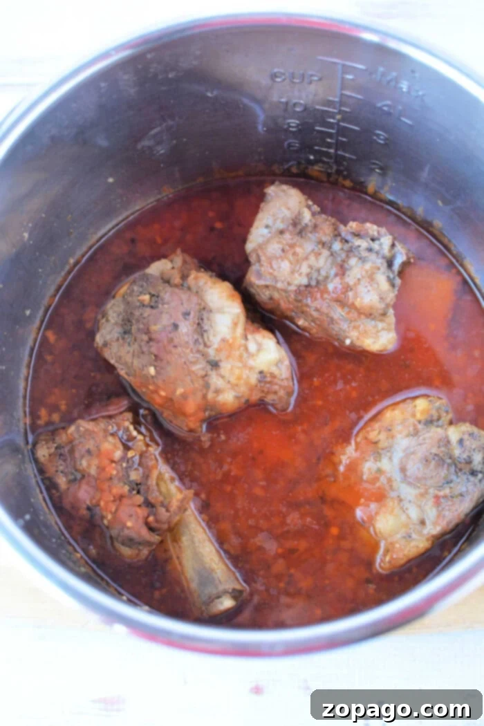 Tender Beef Ribs in the Instant Pot 5 Cooked oxtail in a pot with rich, red broth, hinting at the tenderness achieved by using a pressure cooker. Four pieces are visible alongside aromatic spices and herbs.