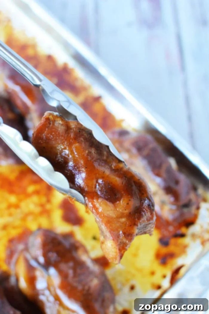 Tongs holding a piece of beautifully glazed boneless pork ribs above a baking tray, ready for the oven, with more succulent, glazed meat waiting beneath.