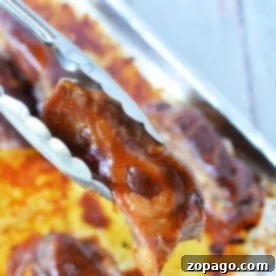 Tongs holding a piece of beautifully glazed boneless pork ribs above a baking tray, ready for the oven, with more succulent, glazed meat waiting beneath.