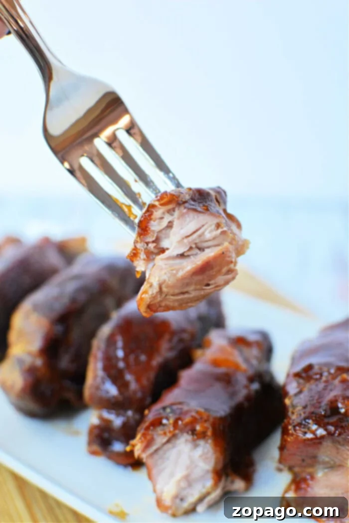 A fork holding a piece of glazed, tender boneless pork ribs above a plate of ribs, showcasing the perfect result of oven cooking.