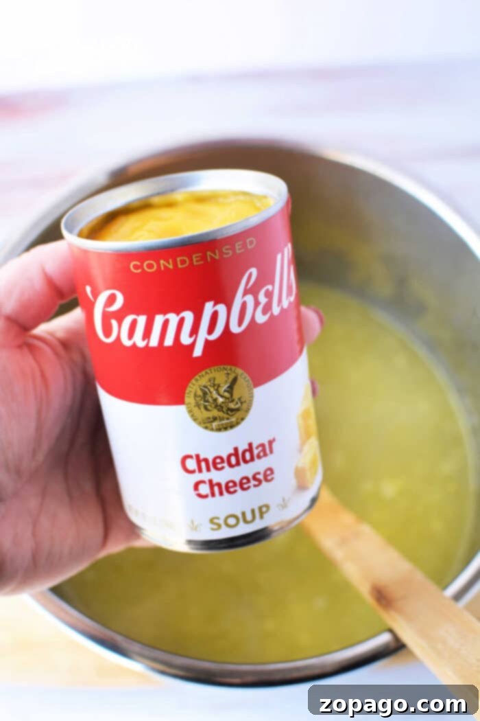 A hand holds a Campbells Cheddar Cheese soup can above a pot containing yellow potato soup with a wooden spoon, perfect for transforming with frozen hash browns in your pressure cooker.