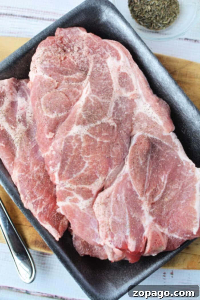 Perfect Pork Steak 3 Raw pork steaks seasoned with pepper on a dark tray, ready for your next Pork Steak Recipe, with a bowl of dried herbs nearby.