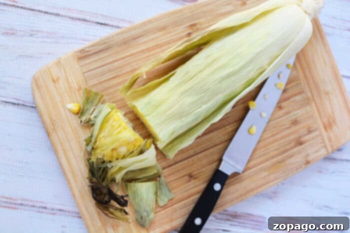 Perfect Oven Roasted Corn on the Cob 6 A partially peeled corn cob with husk rests alongside a knife on a wooden cutting board, ready for an adventure in oven cooking.