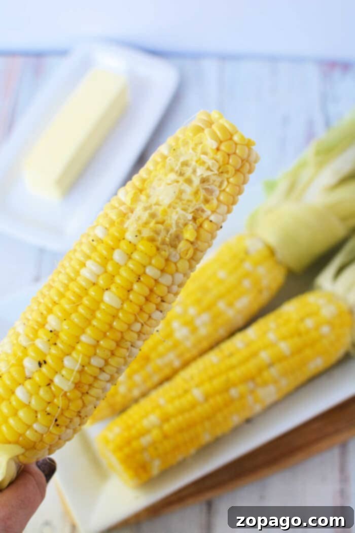 Perfect Oven Roasted Corn on the Cob 7 A close-up shows a buttered ear of corn on the cob, with two unbuttered ones and a stick of butter in the background, capturing the essence of oven-cooked perfection.