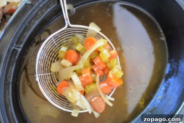 Effortless Slow Cooker Bone Broth 6 A wire ladle lifts diced carrots, celery, and onions above a pot of simmering bone broth, creating a fragrant medley perfect for your slow cooker.