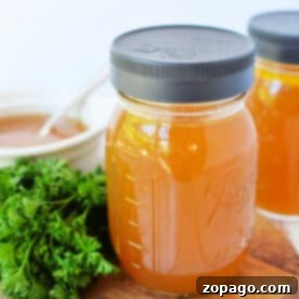 Two mason jars of golden bone broth sit on a wooden board, accompanied by fresh parsley, with a white bowl in the background. Perfect for your favorite slow cooker broth recipe.