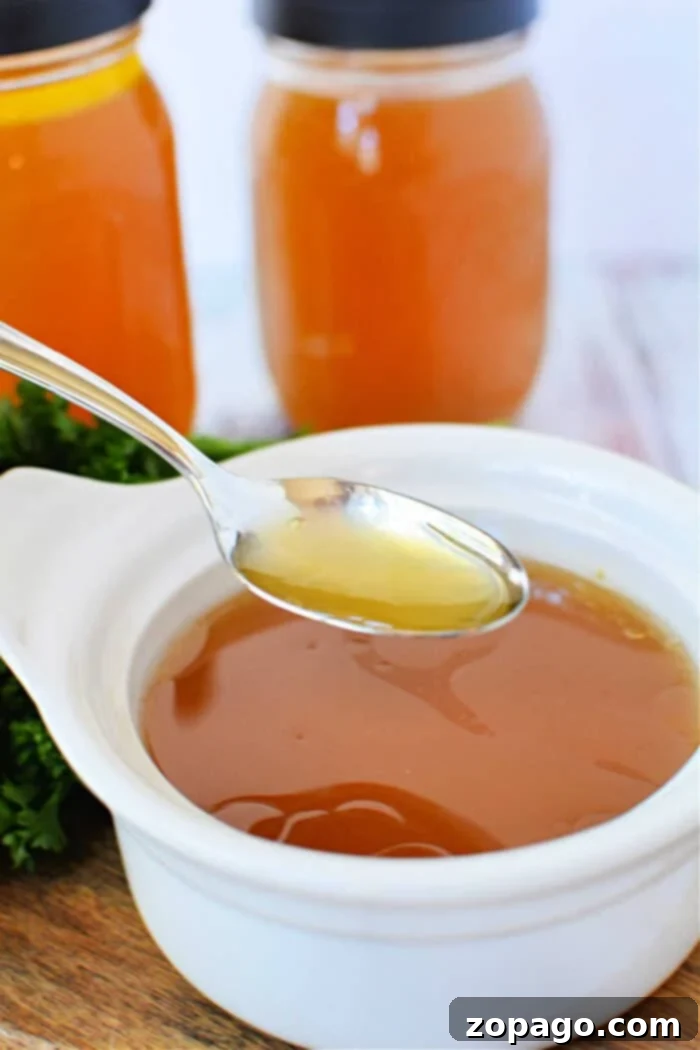 Effortless Slow Cooker Bone Broth 8 A spoon hovers over a white bowl, filled with homemade bone broth made in a slow cooker, with two jars in the background.