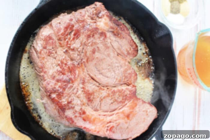 Melt-in-Your-Mouth Boneless Chuck Roast 4 A large boneless chuck roast sizzling in a cast iron skillet, surrounded by seasonings and a measuring cup of broth, ready for the oven.