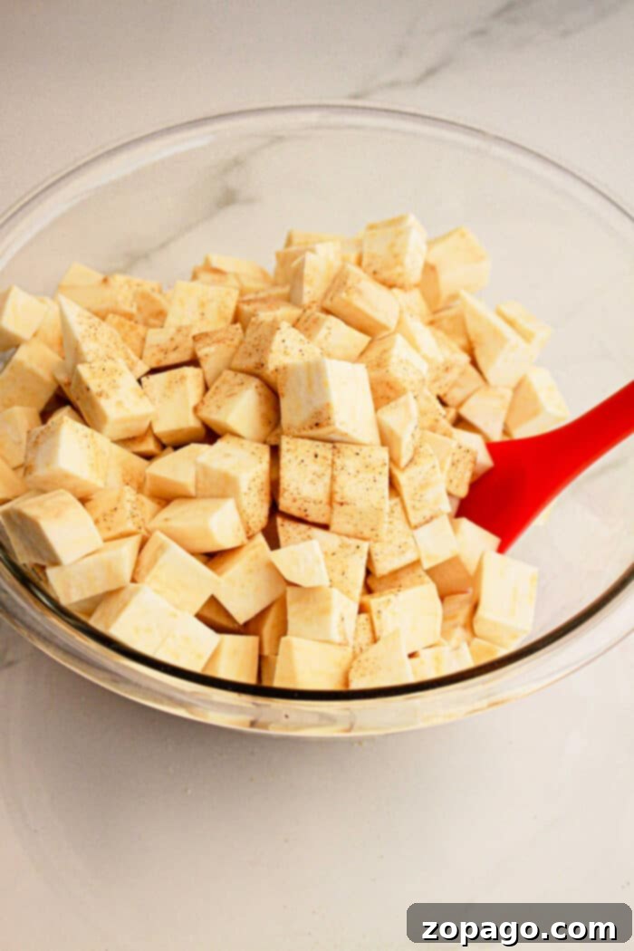 Creamy White Sweet Potato Delight 4 Glass bowl of cubed raw sweet potato with seasoning, stirred by a red spatula on a white countertop—perfect for adding to your favorite White Sweet Potato Recipe.