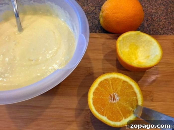 A bowl of vanilla cake batter sits on a wooden board, flanked by a whole orange and a hollowed-out orange half, ready for making grilled orange zest cupcakes.
