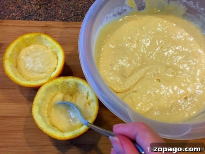 A hand gently spoons vanilla cake batter into hollowed orange halves on a wooden cutting board, preparing them for grilling as a unique BBQ dessert.