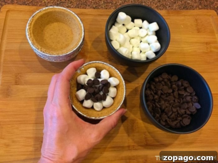 A hand holds a mini pie crust filled with marshmallows and chocolate chips, ready for grilling, with bowls of ingredients nearby for making S'mores pies.