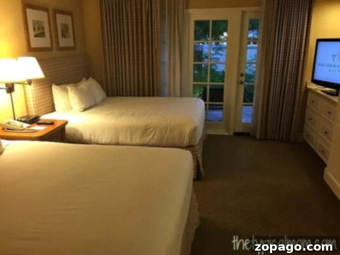 A Look Inside the Arizona Grand Resort 2 The Arizona Grand Resort offers a hotel room featuring two double beds, a nightstand with a lamp, and a dresser. A glass door with curtains opens to a patio with a serene garden view.