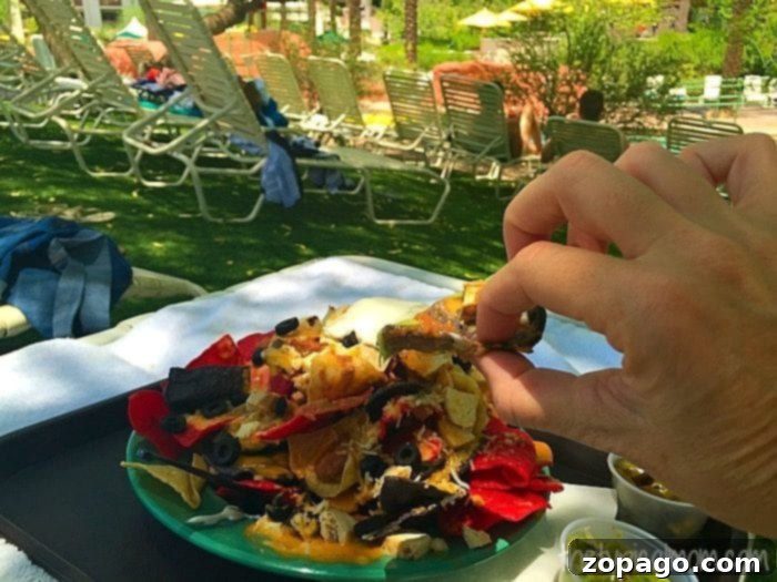 A Look Inside the Arizona Grand Resort 4 A person at the Arizona Grand Resort grabs a loaded nacho chip from a plate, surrounded by lounge chairs and lush green grass, poolside.