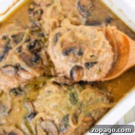 Hamburger steaks in mushroom sauce in a white baking dish, reminiscent of classic Hamburger Steak flavors, with a wooden spoon lifting one piece.