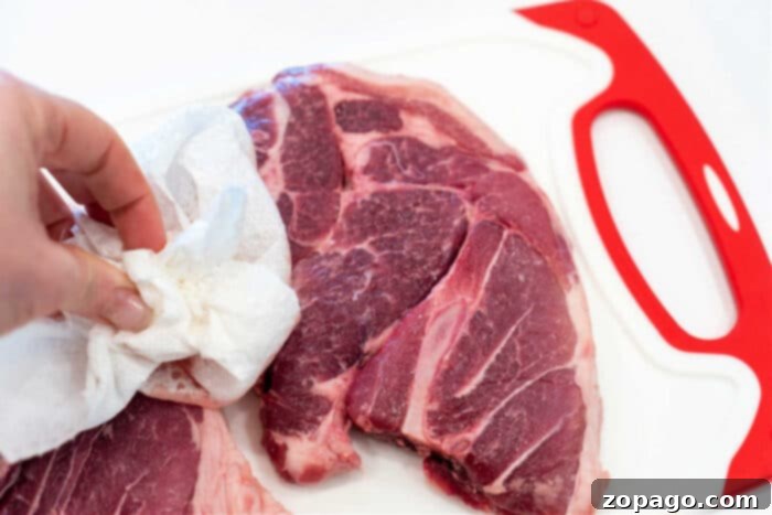 Hearty Pork Shoulder Steak 5 A close-up of a hand dabbing raw pork shoulder steaks with a paper towel on a white cutting board, emphasizing the importance of drying the meat before seasoning.