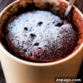 A delightful chocolate mug cake made with cake mix and a hint of mayonnaise for extra moisture, topped with powdered sugar in a beige mug, complete with a spoon inside.